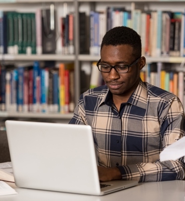 In The Library - Handsome African Male Student With Laptop And Books Working In A High School - University Library - Shallow Depth Of Field